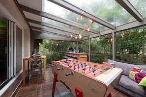 Una sala con paredes de vidrio, una mesa de futbolín, un sofá y una mesa de comedor.