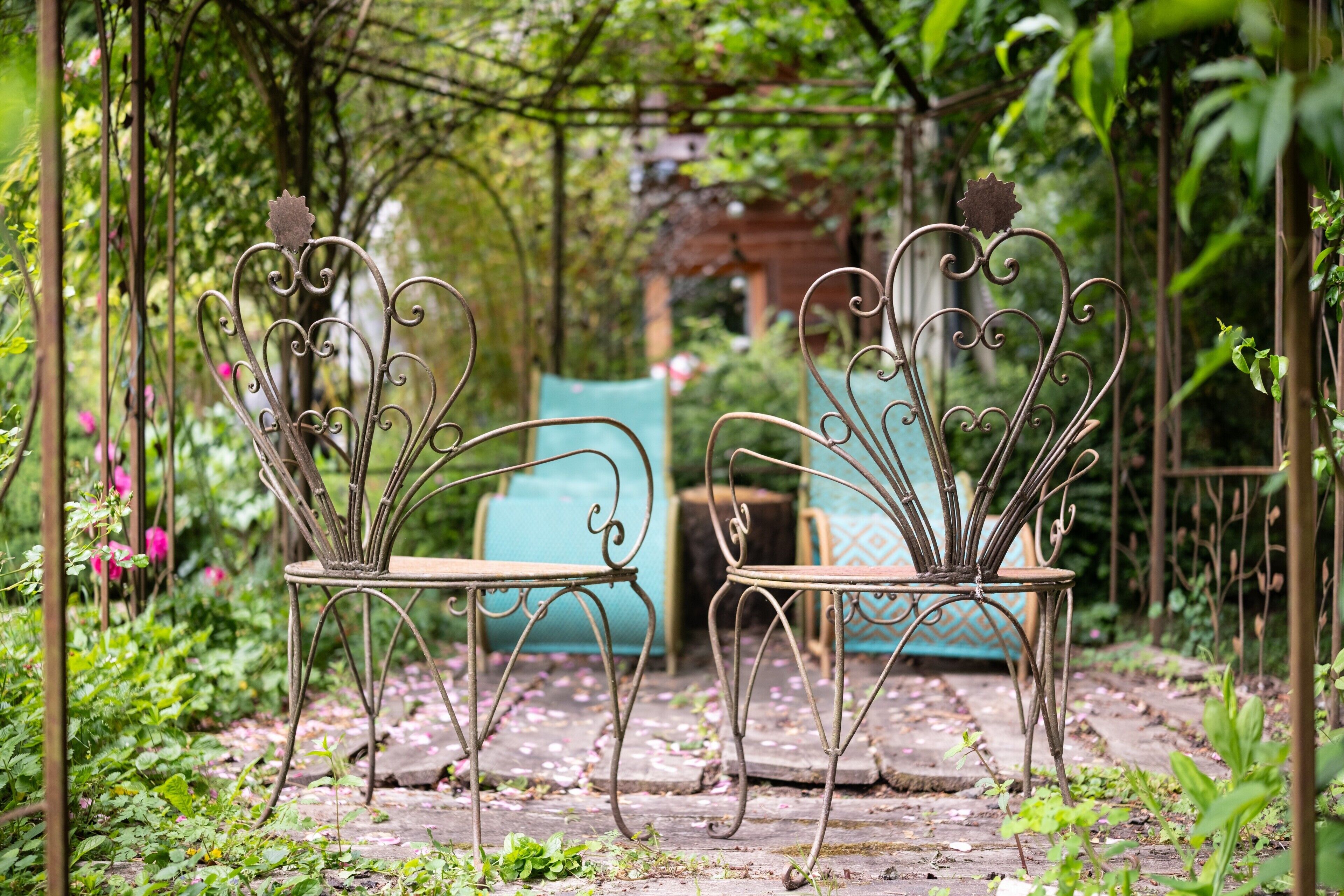 Two metal garden chairs with intricate designs, placed on a stone pathway surrounded by lush greenery and a wooden pergola.