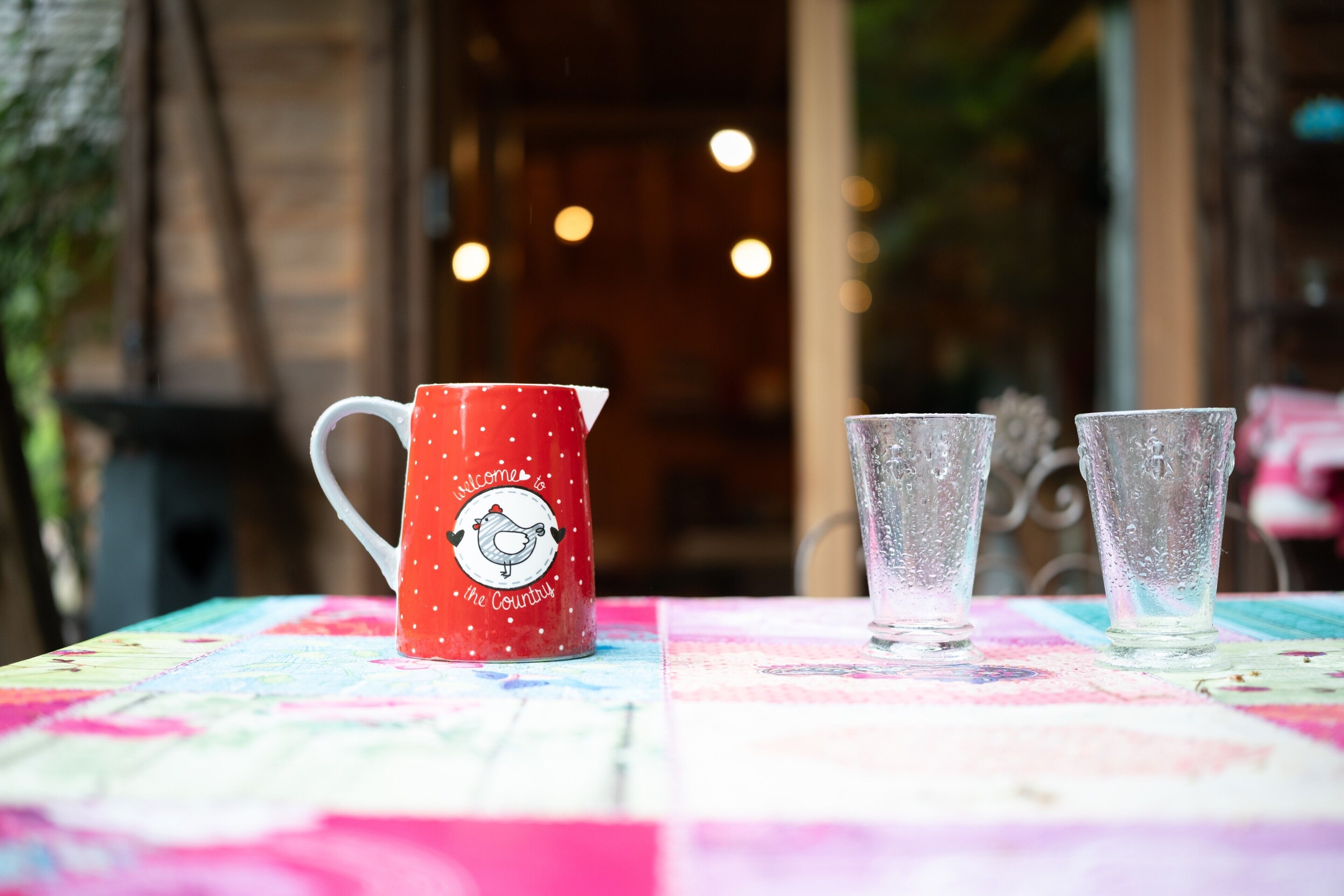 Une table recouverte d’une nappe colorée, un pichet rouge orné d’un motif d’oiseau et deux verres transparents.