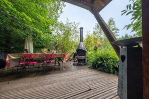 Une terrasse en bois avec une table et des chaises, entourĂ©e de verdure, et un poĂȘle Ă bois.