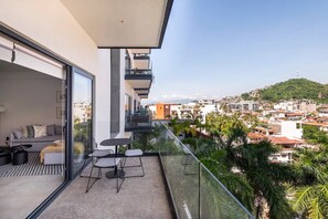 Varanda moderna com sofá, cadeiras e uma mesa, com vista para um horizonte urbano com edifícios e áreas verdes.