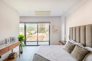 Quarto com uma cama grande, uma mesinha de cabeceira, uma planta em vaso e vista para prédios por uma porta deslizante.