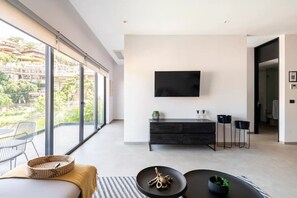 Ein modernes Wohnzimmer mit großem Fenster, einem Flachbildfernseher, einem dunklen Holzschrank und einem kleinen Tisch mit Dekoration.