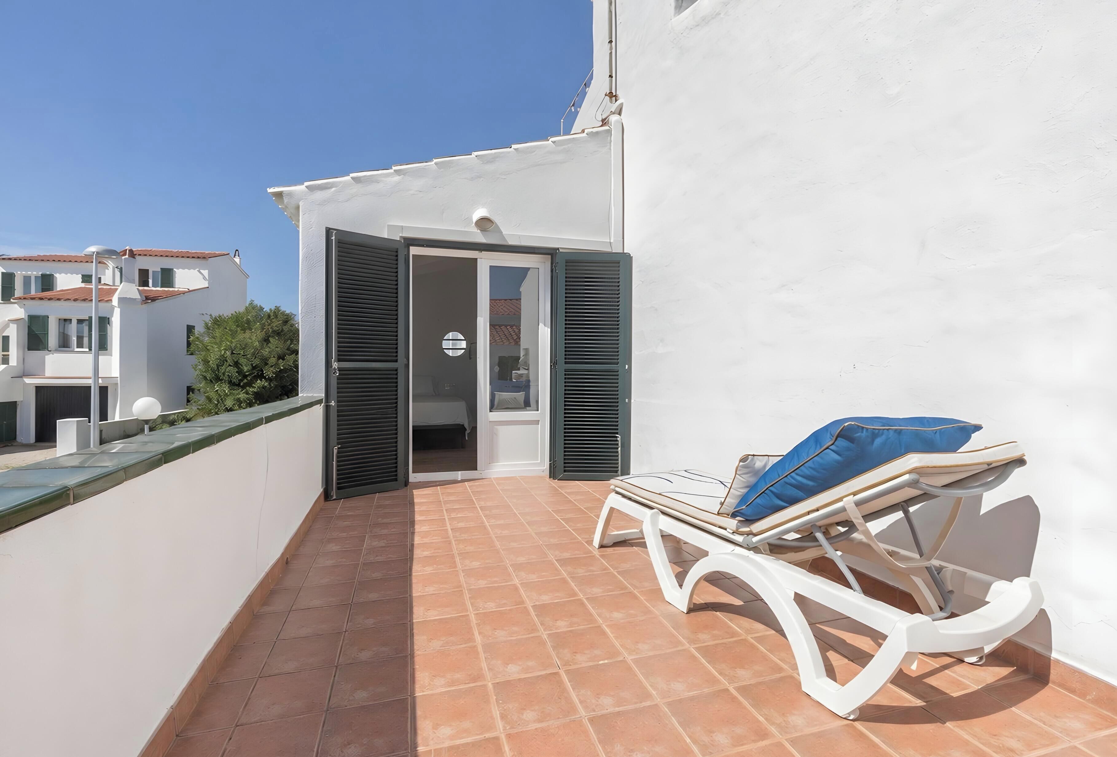 Un balcón con una silla blanca y un cojín azul, una pared blanca y una puerta que da a una habitación interior.