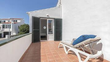 Un balcón con una silla blanca y un cojín azul, una pared blanca y una puerta que da a una habitación interior.