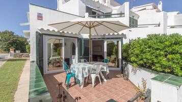 Un bâtiment blanc doté d’un patio abrité comprenant une table et des chaises, un grand parasol et une haie verte.