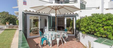A white building with a covered patio area featuring a table and chairs, a large umbrella, and a green hedge.