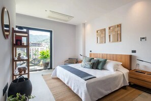 A modern bedroom with a large bed, a wooden nightstand, a bedside table, a bookshelf, and a large window with a view of buildings and mountains. - Loft for rent in the Romantic Zone of Puerto Vallarta (Puerto Vallarta)