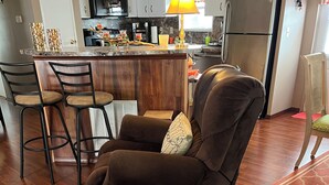 Een woonkamer met een donkerbruine fauteuil, houten vloeren, een keuken op de achtergrond en een eethoek met barstoelen.
