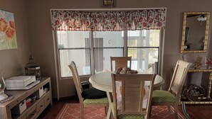 Een eetruimte met een ronde tafel, stoelen, een raam met jaloezieën, een boekenkast en een spiegel.