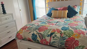 Een bed met een bloemenprint beddegoed, een witte dressoir met lades, een blauw gordijn en een wit hoofdeinde.