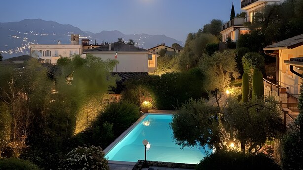 Una piscina rodeada de vegetación y edificios, con montañas al fondo y la luna visible.