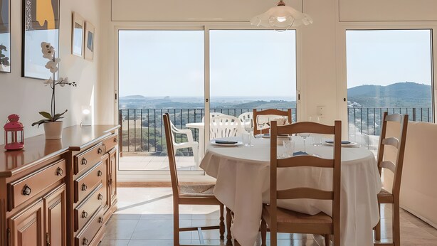 Un comedor con una mesa dispuesta para cuatro, una silla blanca, una lámpara colgante y vistas al paisaje a través de amplios ventanales.