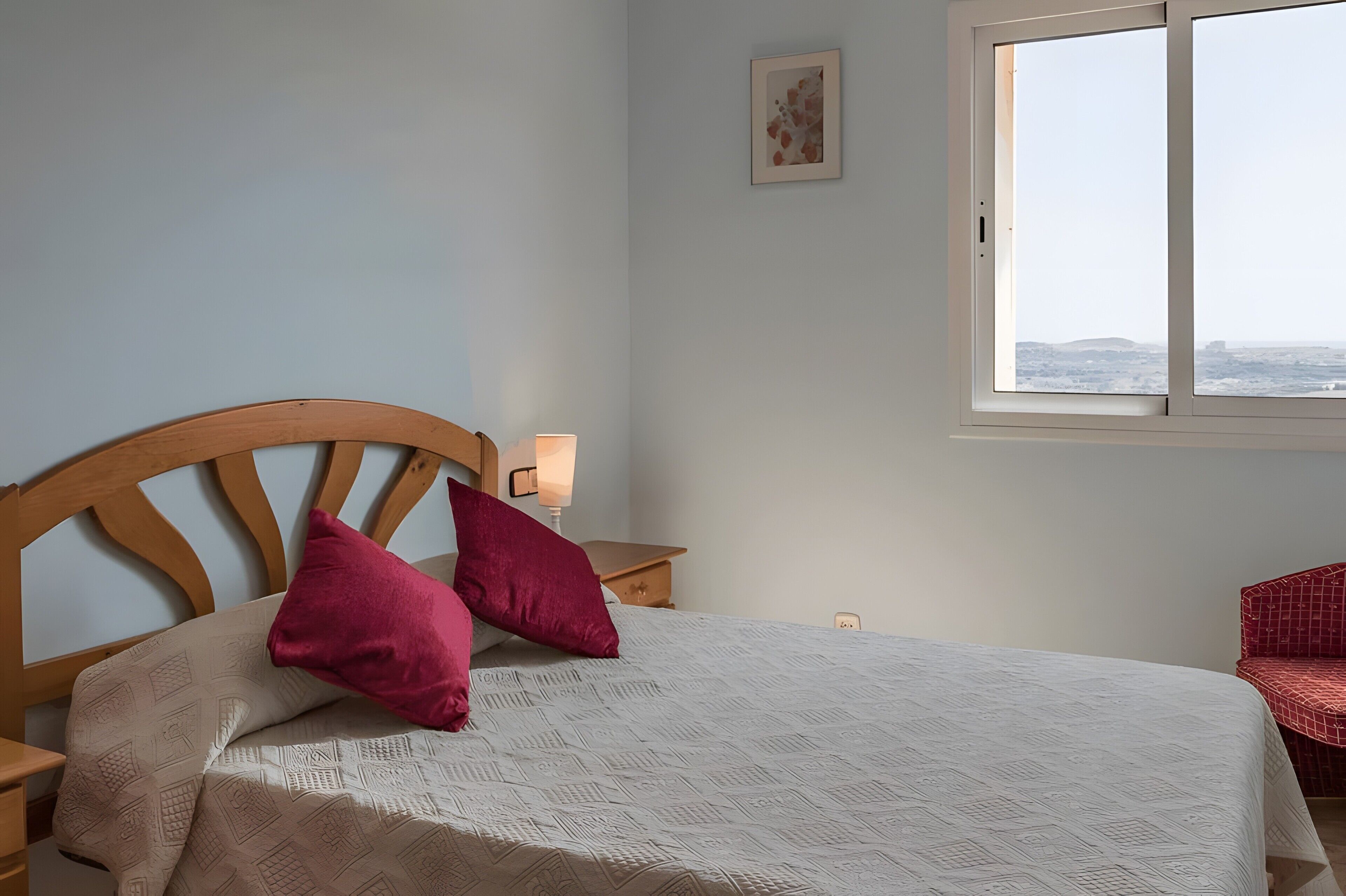 A bed with a headboard, two pillows, a bedside table with a lamp, a wall-mounted picture, and a window with a view.