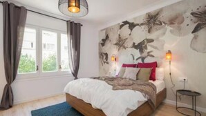 A bedroom with a bed, curtains, a wall mural, two bedside lamps, and a view of greenery outside. - Premium 3-Bedroom Retreat in the Heart of Barcelona (Barcelona)