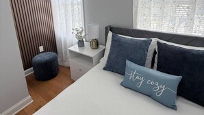 Quarto com cama, duas almofadas, mesa de cabeceira, luminária e janela com cortinas leves.