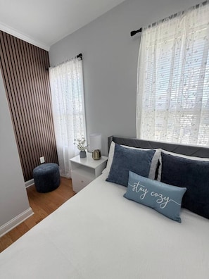 A bedroom with a bed, two pillows, a nightstand, a lamp, and a window with sheer curtains.