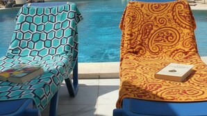 Deux chaises longues, placées au bord d’une piscine, sont dotées de coussins à motifs, et un livre est posé sur l’une d’elles.