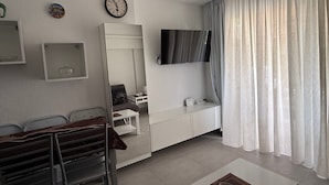 Un salon moderne avec un canapé, une table basse, un téléviseur fixé au mur, une horloge et une fenêtre avec des rideaux.
