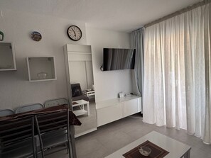 A modern living room with a sofa, a coffee table, a TV mounted on the wall, a clock, and a window with curtains. - Apartment Near the Beach with Ocean View, Just Steps Away (Costa Adeje)