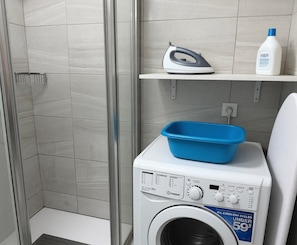 A laundry area with a washing machine, a blue basin, an iron, and a bottle of detergent on a shelf. - Apartment Near the Beach with Ocean View, Just Steps Away (Costa Adeje)