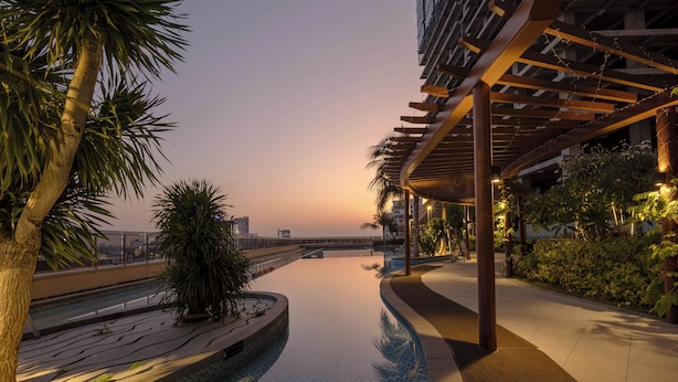 A pool with a tiled edge, palm trees, and a building with a wooden pergola. - Pinnacle Moments Residence (Phnom Penh)