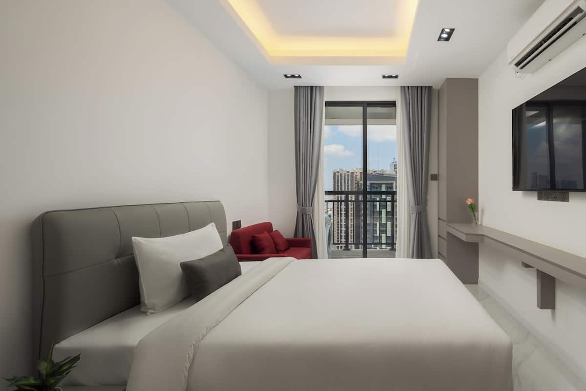 A modern hotel room with a large bed, a red chair, a TV, and a balcony with a city view.