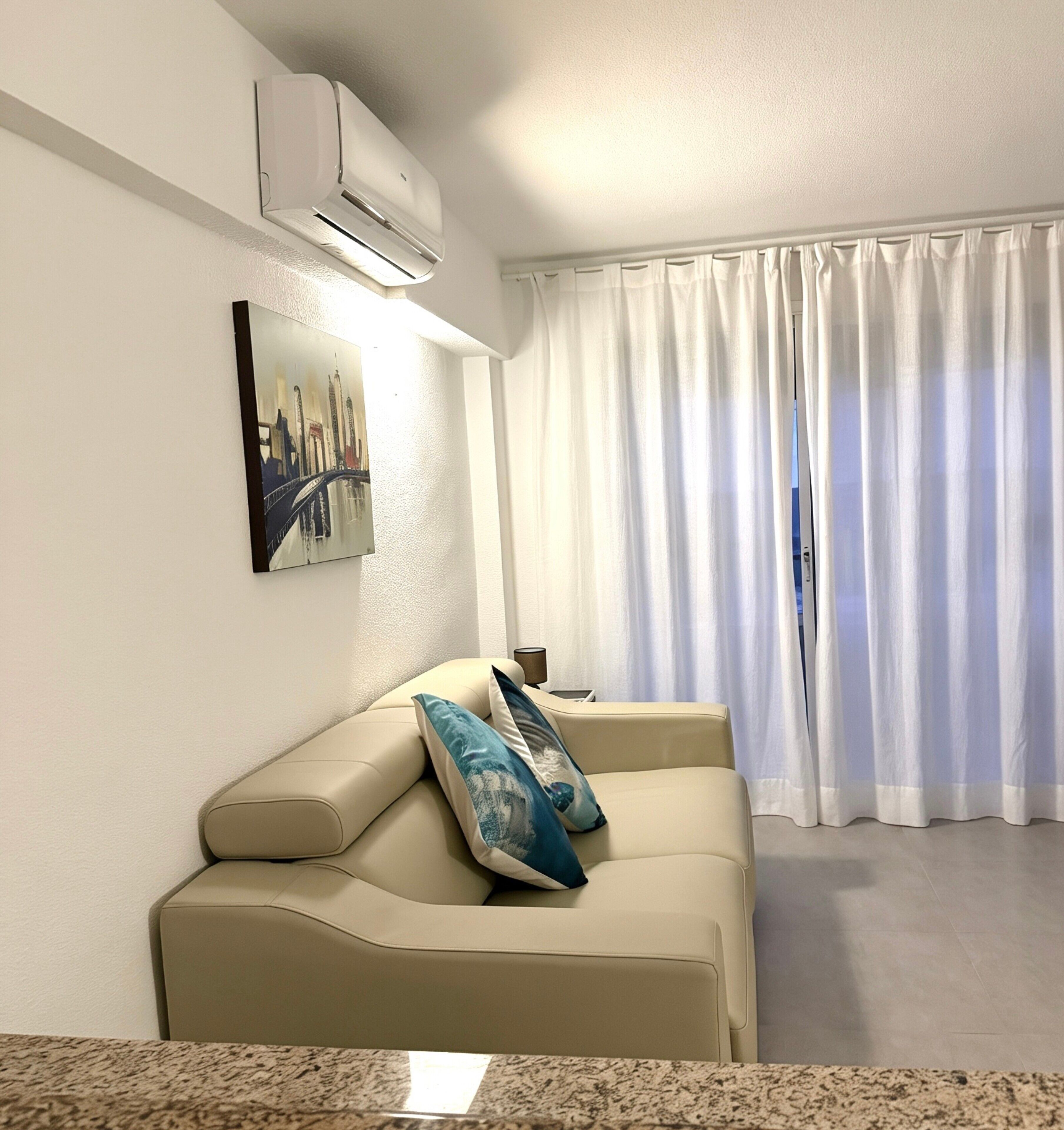 A living room with a beige sofa, a painting of a cityscape, white curtains, and an air conditioning unit.