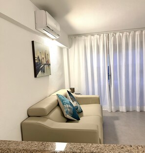 A living room with a beige sofa, a painting of a cityscape, white curtains, and an air conditioning unit. - Sant’Eugenio Basso. Extraordinary Penthouse near the Beaches with Breathtaking Ocean View (San Eugenio Alto)