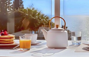 A breakfast table with pancakes, orange juice, a teapot, and glasses, overlooking a scenic ocean view. - Sant’Eugenio Basso. Extraordinary Penthouse near the Beaches with Breathtaking Ocean View (San Eugenio Alto)