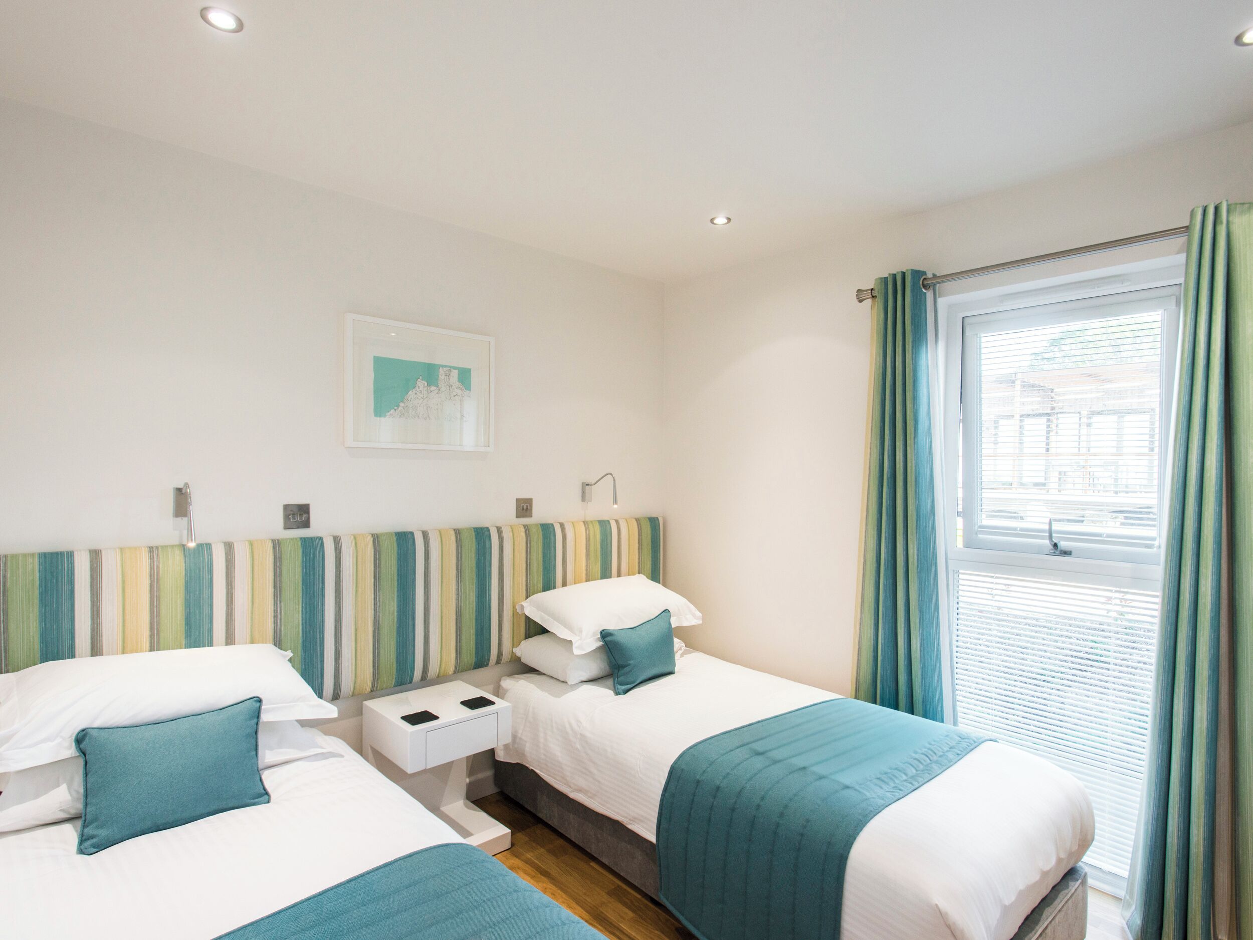A hotel room with two beds, a striped headboard, a window with curtains, and a small bedside table with a lamp.
