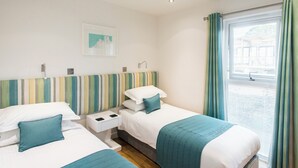 A hotel room with two beds, a striped headboard, a window with curtains, and a small bedside table with a lamp. - Barbara (Redruth)