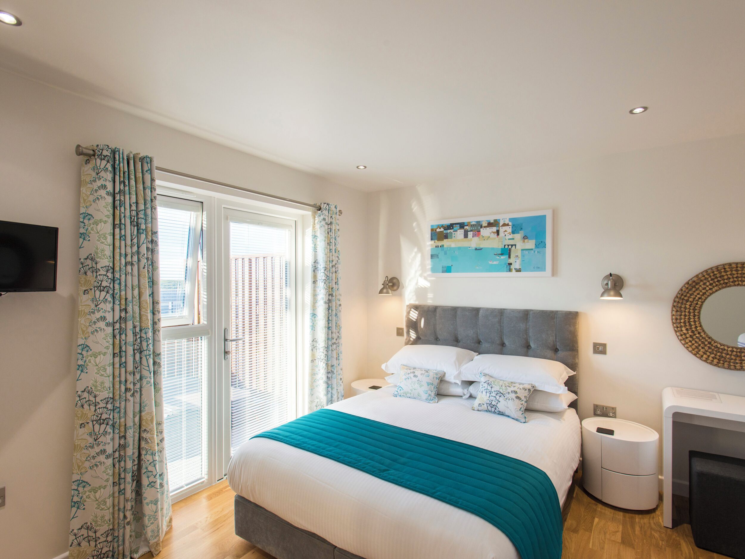 A bedroom with a bed, curtains, a painting, a bedside lamp, a mirror, and a nightstand.