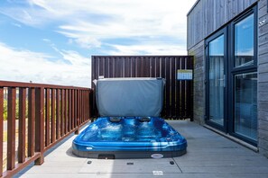 A hot tub on a wooden deck with a wooden fence and a building with large windows. - Barbara (Redruth)