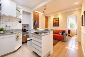 A modern kitchen with a built-in island, a dining area with a red sofa, and a large window providing natural light. - Comfort in Ipanema Near the Beach (Rio de Janeiro)
