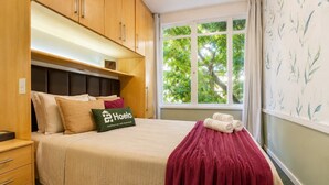 A hotel room with a bed, a nightstand, a window with a view of trees, and a wall with a leaf pattern. - Comfort in Ipanema Near the Beach (Rio de Janeiro)