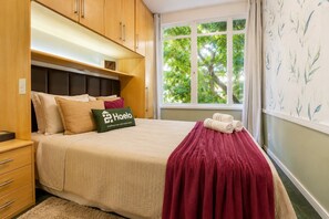 A hotel room with a bed, a nightstand, a window with a view of trees, and a wall with a leaf pattern. - Comfort in Ipanema Near the Beach (Rio de Janeiro)