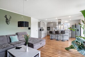 En moderne stue med en grå sofa, et sofabord og en vase med blomster. I baggrunden ses et køkken med et spiseområde.