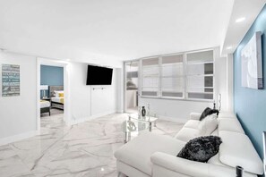 A modern living room with a white sofa, a glass coffee table, and a flat-screen TV mounted on the wall. - Bright & Renovated 2BR by the Ocean (Hollywood)