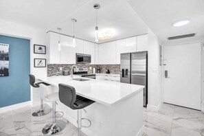 A modern kitchen with white cabinetry, a central island, stainless steel appliances, and pendant lighting. - Bright & Renovated 2BR by the Ocean (Hollywood)