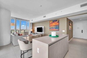 A modern hotel room with a large window, a flat-screen TV, a sofa, a dining area, and a painting on the wall. - Luxury Ocean View 2 BR unit (Hollywood)
