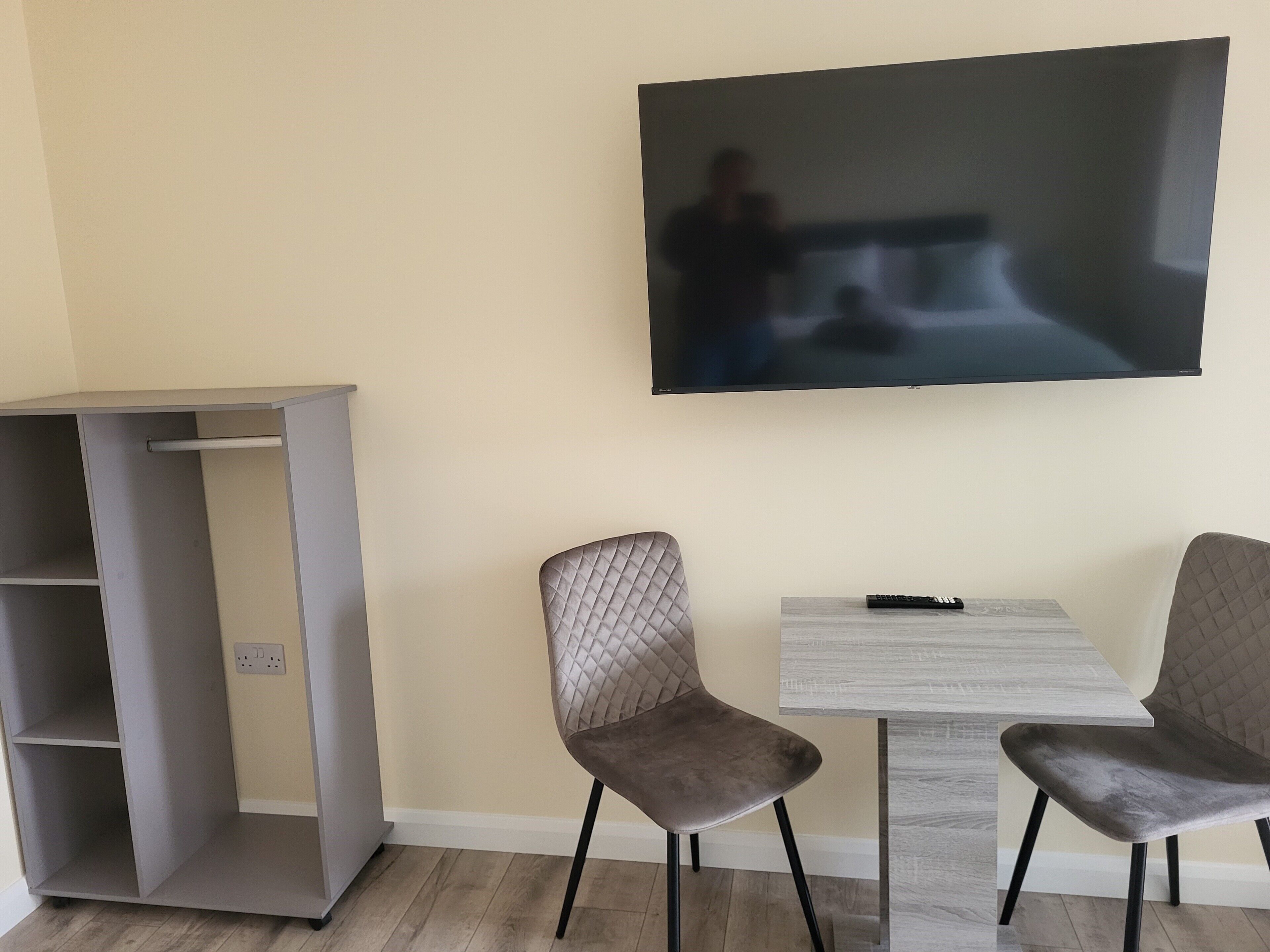 A room with a TV mounted on the wall, two chairs, a small table, and a wardrobe.