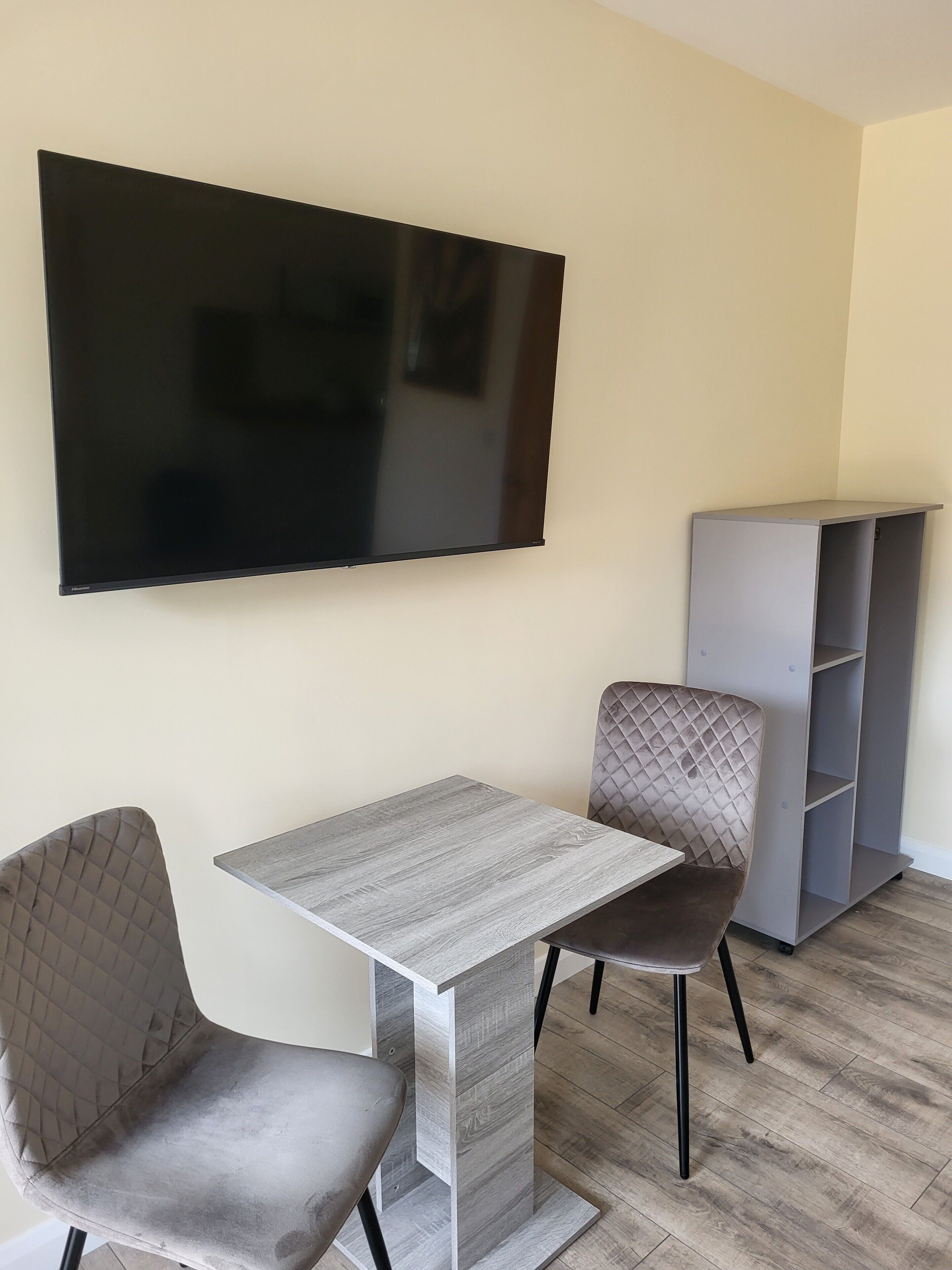Una habitación con una televisión de pantalla plana montada en la pared, un sillón tapizado en gris, una mesa gris y una estantería gris.