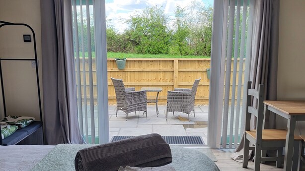 A bedroom with a bed, a small table, and a chair. There is a view of a patio with a wooden fence and outdoor furniture. - The Old Barn (Redditch)