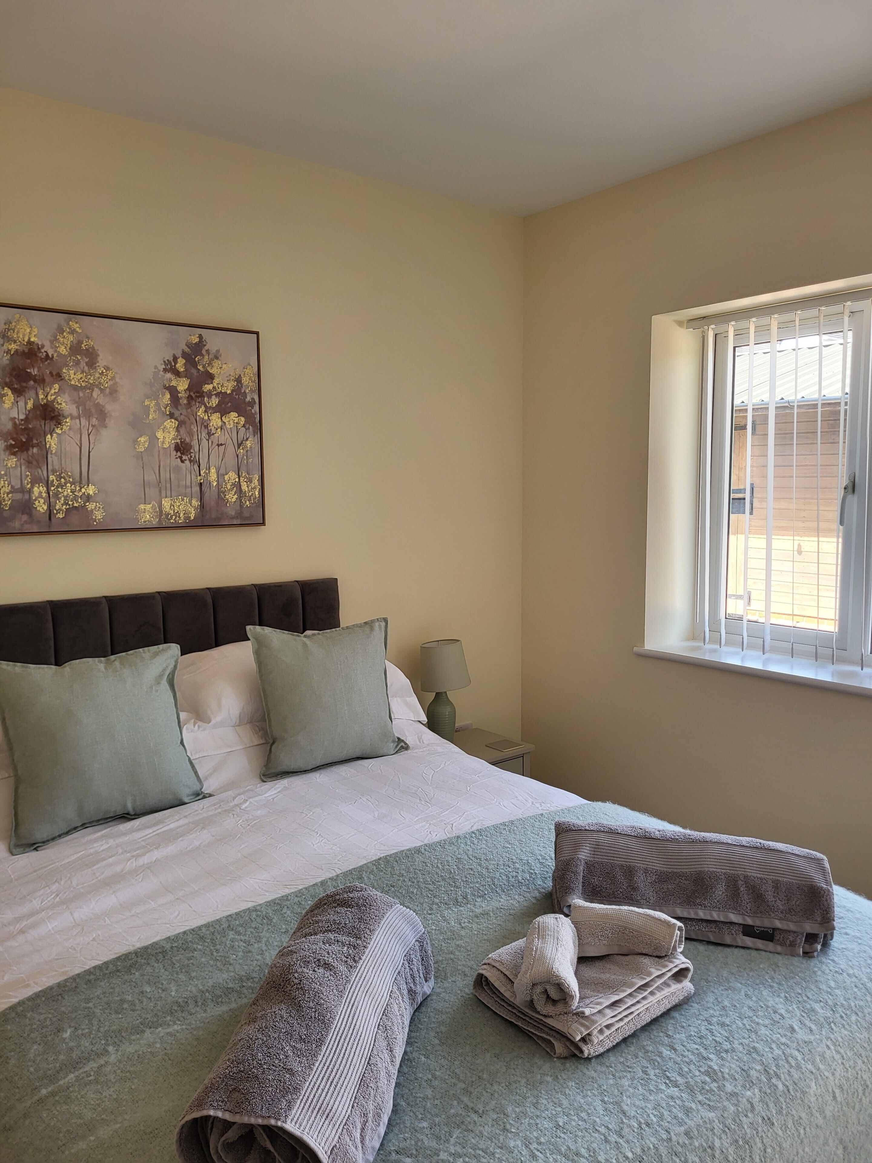 A neatly made bed with pillows, a bedside lamp, and a window with blinds.