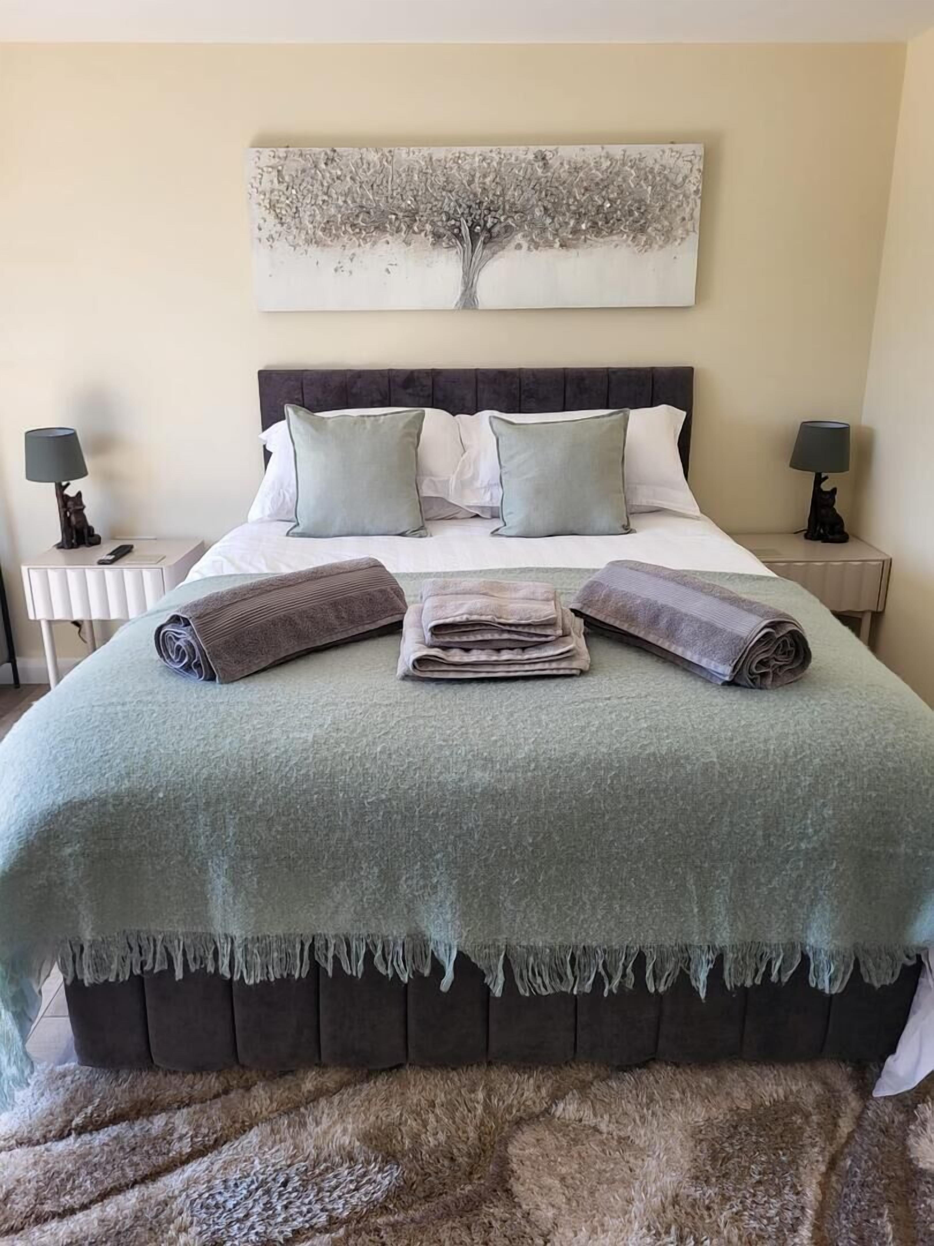 A neatly made bed with a green blanket, pillows, and rolled towels. There is a tree artwork above the bed and two bedside lamps with cat figurines.