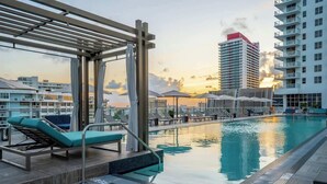 Une piscine sur le toit, dotée de chaises longues, d’une pergola et offrant une vue sur les bâtiments de la ville au coucher du soleil.
