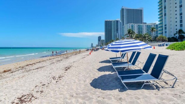 A beach with lounge chairs and a striped umbrella, a clear sky, and high-rise buildings in the background. - Beautiful renovated 2BR unit ocean (Hollywood)