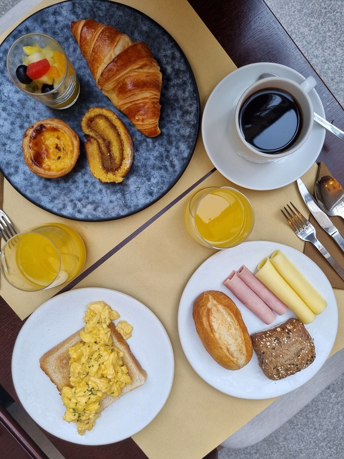 Disposição para o pequeno-almoço com croissants, ovos mexidos, fiambre, queijo e sumo.