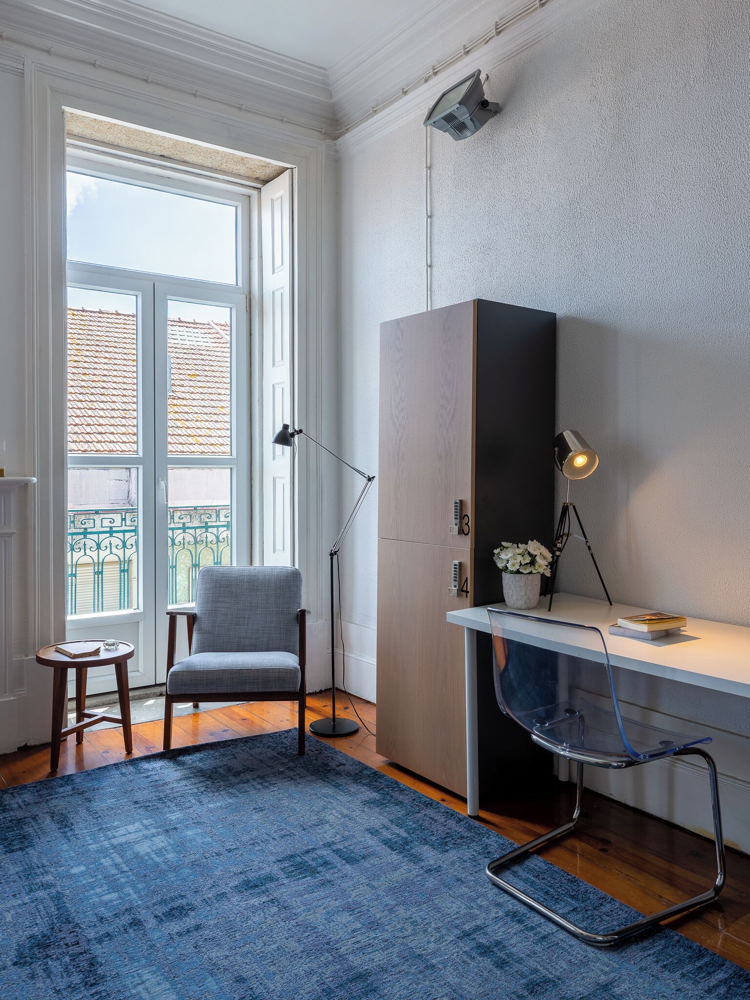 Quarto com escrivaninha, cadeira e uma grande janela com vista para edifícios.